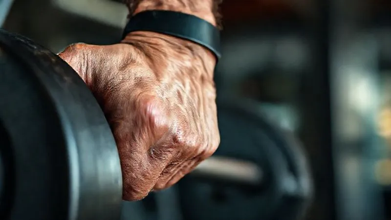 photorealistic, professional photography, 8K, cinematic lighting, sharp focus, depth of field, shot on a high-end DSLR. A detailed, close-up shot of a man's hand in his late 40s, calloused but strong, gripping a heavy barbell. The focus is on the textured metal and the tension in his forearm, conveying the effort and dedication required for strength training. The background is a blurred gym setting, emphasizing the personal struggle and triumph.