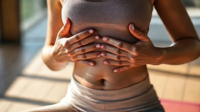A photorealistic, professional photography shot of a person in a serene setting, perhaps outdoors or in a minimalist yoga studio, demonstrating diaphragmatic breathing. Their hands are gently placed on their abdomen, which is visibly rising with a deep, calm inhale. Cinematic lighting highlights their relaxed posture and peaceful expression. Sharp focus on the breathing motion, with a soft depth of field blurring the background. Shot on a high-end DSLR, 8K, conveying tranquility and mindful breathing.