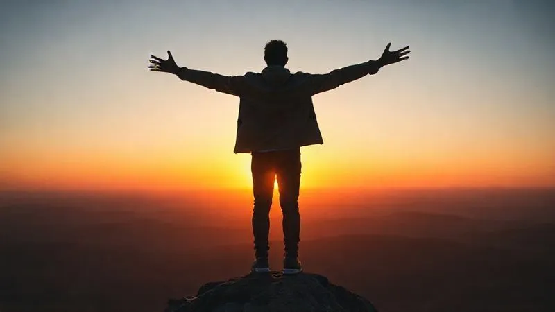 A photorealistic image of a person standing confidently at the edge of a vast, open landscape, with their back to the camera, arms outstretched toward a vibrant sunrise. The silhouette suggests freedom and a hopeful future. Professional photography, 8K, cinematic lighting, sharp focus, depth of field, shot on a high-end DSLR.